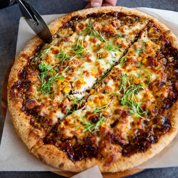 A pizza with melted cheese, corn, chicken, and green sprouts being sliced with a pizza cutter.